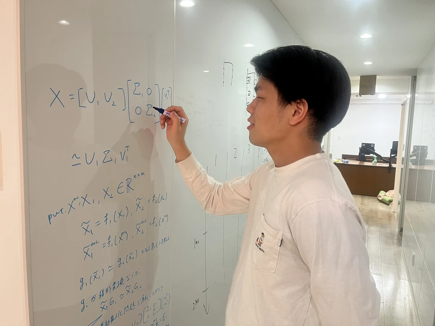 オフィスのホワイトボードに行列分解の数式を書きながら検討する齊藤